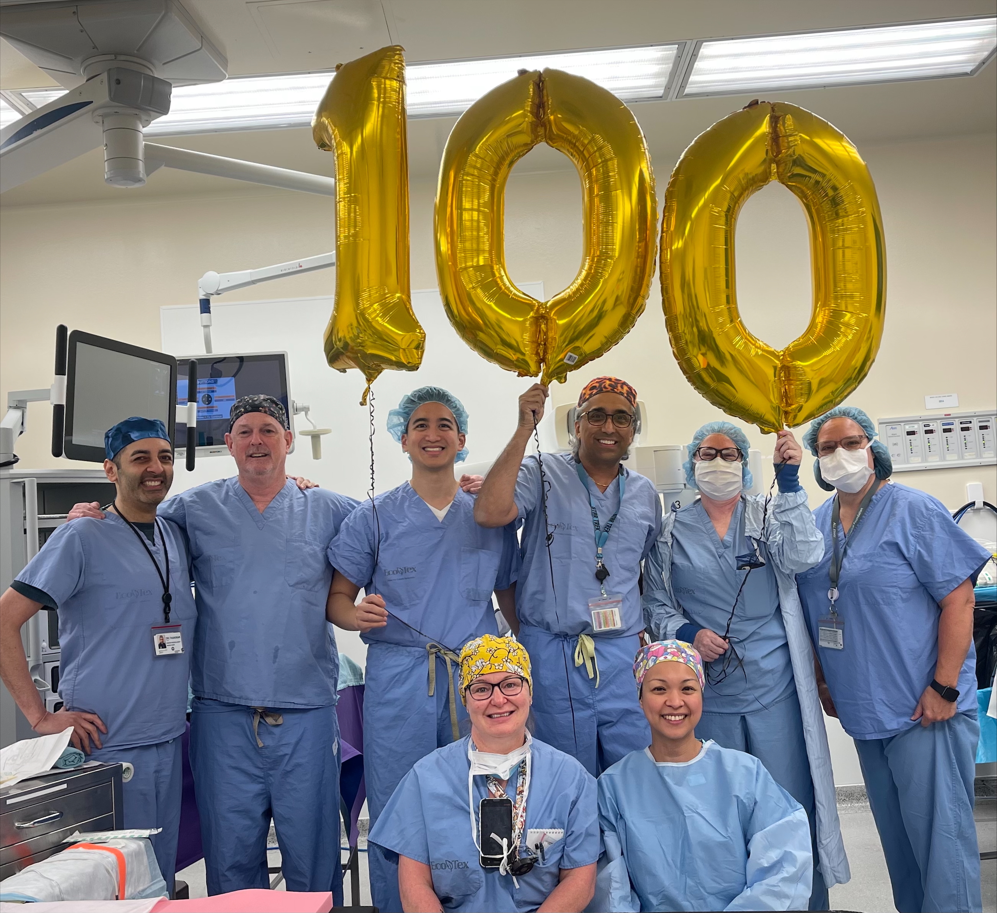 Surgical team standing in front gold foil balloons that read "100."