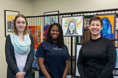 Three women smiling with art pieces on the wall behind them
